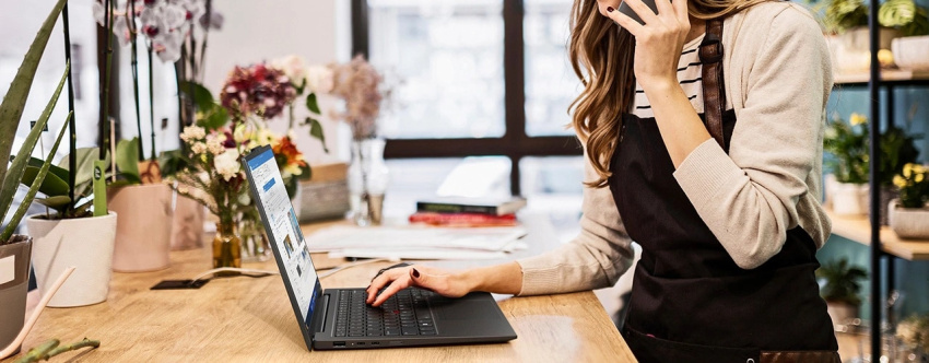 女士坐在植物環繞的書桌旁，一邊使用智能手機，右手一邊在 Lenovo ThinkPad X1 Carbon Gen 12 筆記簿型電腦上打字。